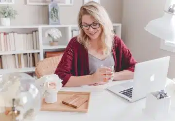 woman smiling holding glass mug sitting beside table with MacBook